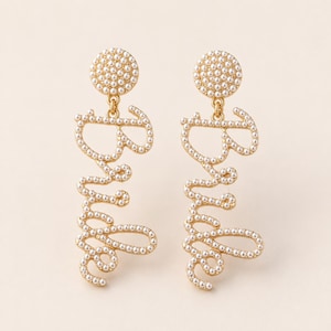 May include: A pair of gold-toned drop earrings featuring the word Bride in cursive script. Each earring is embellished with small white faux pearls along the lettering and a circular stud top also covered in white faux pearls.