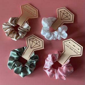 May include: Four satin scrunchies in different colors, beige, white, green, and pink, with a diamond-shaped tag attached to each. The tag has the text "To have and to hold your hair back" in a gold script font.