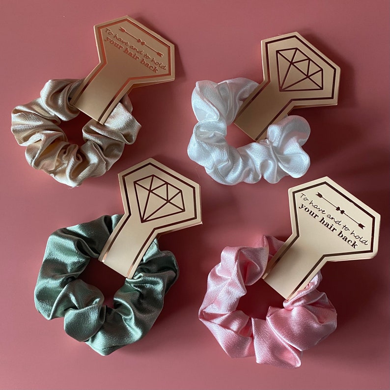May include: Four satin scrunchies in different colors, beige, white, green, and pink, with a diamond-shaped tag attached to each. The tag has the text "To have and to hold your hair back" in a gold script font.