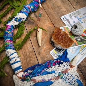 May include: A handmade wand wrapped in blue and white fabric with lace, beads, and a bone charm. The wand is lying on a wooden surface with a tarot card, a crystal, and a fern.