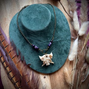 May include: A unique necklace featuring a bone pendant suspended from a bronze chain. The necklace includes purple beads and is displayed on a green velvet bust. The background includes feathers and dried plants.