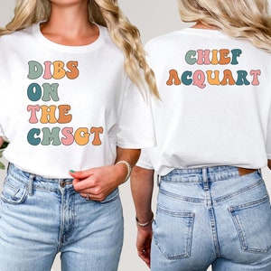 May include: White t-shirt with the text "DIBS ON THE CMSGT" in colorful retro font on the front. The back of the shirt features the text "CHIEF ACQUART" in a similar style. The shirt is worn with blue jeans.