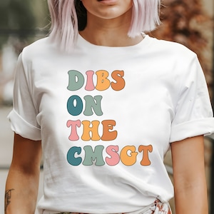May include: White t-shirt with the text "DIBS ON THE CMSGT" in colorful retro-style lettering.