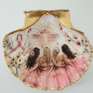 Peut inclure: Un coquillage Saint-Jacques décoratif avec un bord peint en or, présentant une illustration imprimée de quatre personnes en robes roses debout ensemble, un ruban de sensibilisation rose, un soleil et des motifs floraux sur fond blanc.