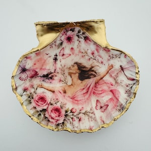 Peut inclure: Un coquillage Saint-Jacques décoratif avec un bord peint en or et une illustration détaillée d'une personne aux bras étendus. La scène comprend des roses roses, des papillons, un phare et une libellule sur un fond blanc doux.