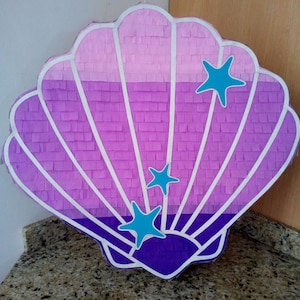 May include: A purple and pink seashell shaped pinata with white trim and blue star decorations.