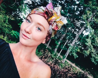 May include: A woman with a light complexion and blue eyes is wearing a colorful patterned headscarf. She is smiling and looking at the camera. The background is a blurred image of green trees.