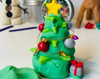 Op de afbeelding: Een groene kerstboom van klei versierd met een gele ster, rode kerstballen, zilveren en gouden kralen en witte en roze lampjes. De boom is omringd door andere klei-creaties, waaronder een groene kleibol en een grijze cadeaudoos. De tekst "Maak je eigen kerstboom" staat onder de boom.