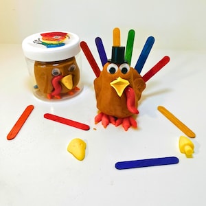 May include: Thanksgiving-themed craft kit featuring a brown clay turkey with colorful popsicle stick feathers, googly eyes, and a tiny black hat. A jar of brown clay with a turkey face is also present, along with additional craft supplies.