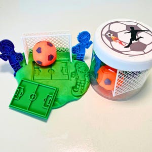May include: A set of soccer-themed play dough tools, including a goal, a soccer ball, and two figures of players. The tools are displayed on a green play dough surface. A clear jar with a white lid contains two orange soccer balls and a white net.