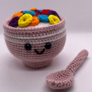 Puede incluir: Un cuenco de crochet rosa lleno de bucles de cereales de crochet de colores y una cuchara de crochet rosa a juego. El cuenco tiene un borde de crochet blanco y una cara sonriente con ojos negros.