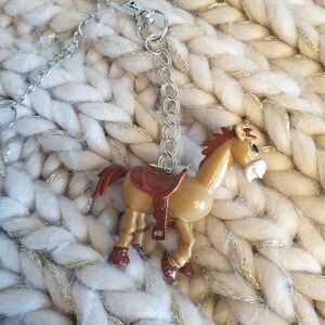 May include: A brown plastic toy horse with a saddle and a silver chain attached to a silver clasp. The horse is on a white and gold knitted background.