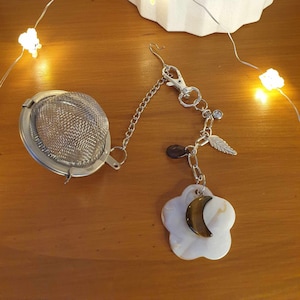 May include: A silver tea infuser with a chain and a charm attached. The charm is a white flower with a crescent moon made of tiger's eye stone.