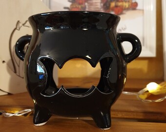 May include: Black ceramic cauldron with a bat-shaped cutout design. The cauldron has three legs and two handles.