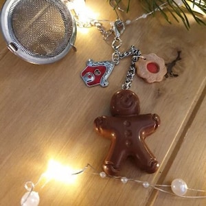 May include: A silver tea infuser with a gingerbread man, a house, and a star charm. The charms are attached to a silver chain. The infuser is on a wooden surface with white string lights.