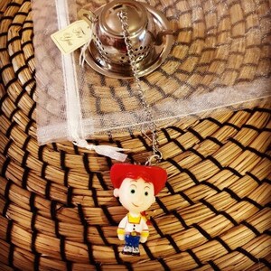 May include: A silver tea infuser with a chain and a red and yellow plastic toy character of a girl with a red cowboy hat. The toy character is attached to the chain of the tea infuser. The tea infuser and toy character are on a woven straw mat.