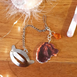 May include: A silver hair clip with a chain and a charm featuring a brown and white dog head. The chain also has an amber-colored bead.