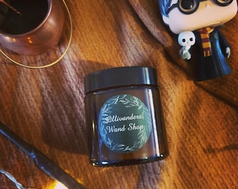 Peut inclure: Une bougie dans un bocal en verre brun avec un couvercle noir et une étiquette verte et blanche qui dit "Ollivanders' Wand Shop".