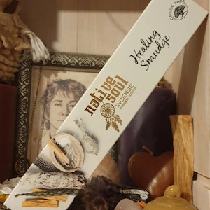 Peut inclure: Une boîte d'encens Native Soul Healing Smudge. La boîte est blanche avec des lettres dorées et un motif de capteur de rêves et de bâtonnets d'encens. La boîte est posée sur une table avec d'autres objets.