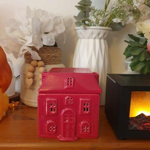 Peut inclure: Un chauffe-cire en céramique rouge en forme de maison avec une porte et des fenêtres blanches. Le chauffe-cire est posé sur une surface en bois.