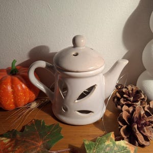 Peut inclure: Un photophore en céramique blanc en forme de théière avec des découpes en forme de feuilles. La théière a un couvercle avec un petit bouton et un bec. Une citrouille orange, des pommes de pin et des feuilles d'automne sont disposées autour de la théière.