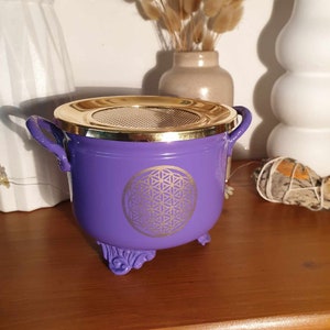 May include: Purple metal incense burner with a gold lid and a gold flower of life design. The burner has three decorative feet.