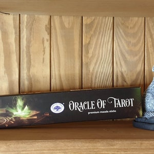 May include: A black box of "Oracle of Tarot" premium masala sticks is displayed on a wooden shelf. The box features a mystical design with a glowing orb and a pentagram. A grey wolf statue is to the right.