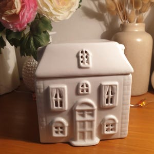 Peut inclure: Un récipient blanc en céramique en forme de maison avec un toit en pente, des fenêtres et une porte d'entrée. Le récipient est conçu avec une surface détaillée et texturée. La maison est posée sur une surface en bois.