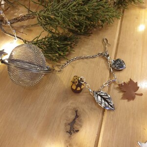 May include: A silver tea infuser with a chain and charms. The charms include a heart, a leaf, a small acorn, and a brown maple leaf.
