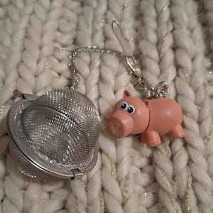 Peut inclure: Infuseur à thé en acier inoxydable avec une chaîne et un charm en forme de cochon rose. L'infuseur est en forme de boule en maille. Le charm en forme de cochon a des yeux noirs et est rose clair. Fond en tissu tricoté crème.
