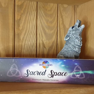Peut inclure: Une boîte d'encens Masala Premium Sacred Space avec un symbole de triskèle et un motif de ciel étoilé. La boîte est posée sur une étagère en bois avec une figurine de loup gris en train de hurler à la lune.