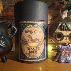 Peut inclure: Une boîte à thé cylindrique noire avec une étiquette intitulée Trelawney's Divination Tea, Nessie's Brew Scottish Breakfast. L'étiquette montre une tasse de thé avec une créature à l'intérieur, entourée de motifs célestes dorés.