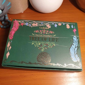 May include: A green box with gold lettering and illustrations of feathers and seashells. The box contains Goloka Sacred Smudges Tree of Life resin incense with cups.