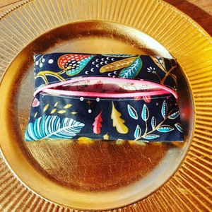 May include: A small, rectangular fabric pouch with a floral pattern in teal, red, yellow, and white on a navy blue background. A bright pink elastic band secures the pouch. The pouch is on a gold-colored plate.