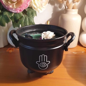 Peut inclure: Bougie noire en forme de chaudron avec un motif de main de Hamsa blanc et une bougie verte à l'intérieur. La bougie est surmontée d'une fleur blanche.