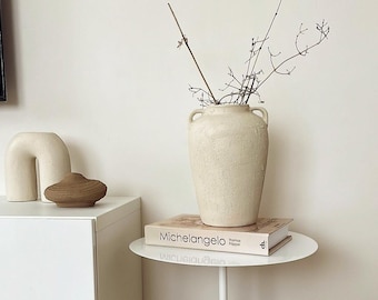 May include: A white side table with a round top and a round, beige base. A book titled "Michelangelo" by Thomas  Hopper is on top of the table, and a white ceramic vase with dried branches is on top of the book.