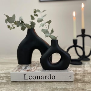 May include: Two black ceramic vases with unique shapes, one with a rounded arch and the other with a circular cutout, holding sprigs of eucalyptus on a book titled "Leonardo".
