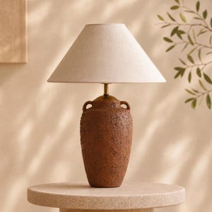 May include: A table lamp featuring a textured, dark brown, amphora-shaped base with two small handles at the neck. The lamp is topped with a conical, off-white fabric shade and includes a simple brass-colored metal stem connecting the base to the shade.