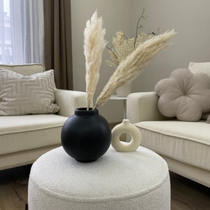 May include: A black spherical vase holds tall, beige pampas grass. Beside it sits a small, cream-colored vase with a circular opening. The vases are on a white, textured ottoman, with cream-colored armchairs and decorative pillows in the background.
