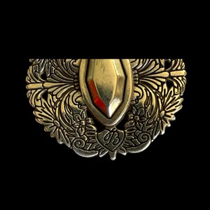 May include: An ornate, antique-style metal brooch with a central gold-coloured shield shape. The brooch features intricate floral and leaf patterns, with a heart-shaped emblem bearing the initials "UB". The background is black.