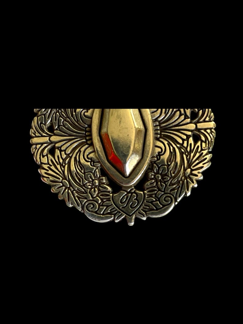 May include: An ornate, antique-style metal brooch with a central gold-coloured shield shape. The brooch features intricate floral and leaf patterns, with a heart-shaped emblem bearing the initials "UB". The background is black.
