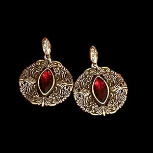 May include: A pair of gold-coloured earrings with a detailed filigree design. Each earring has a central, oval-shaped, deep red gemstone. The earrings feature a floral and scrollwork pattern around the gemstone.