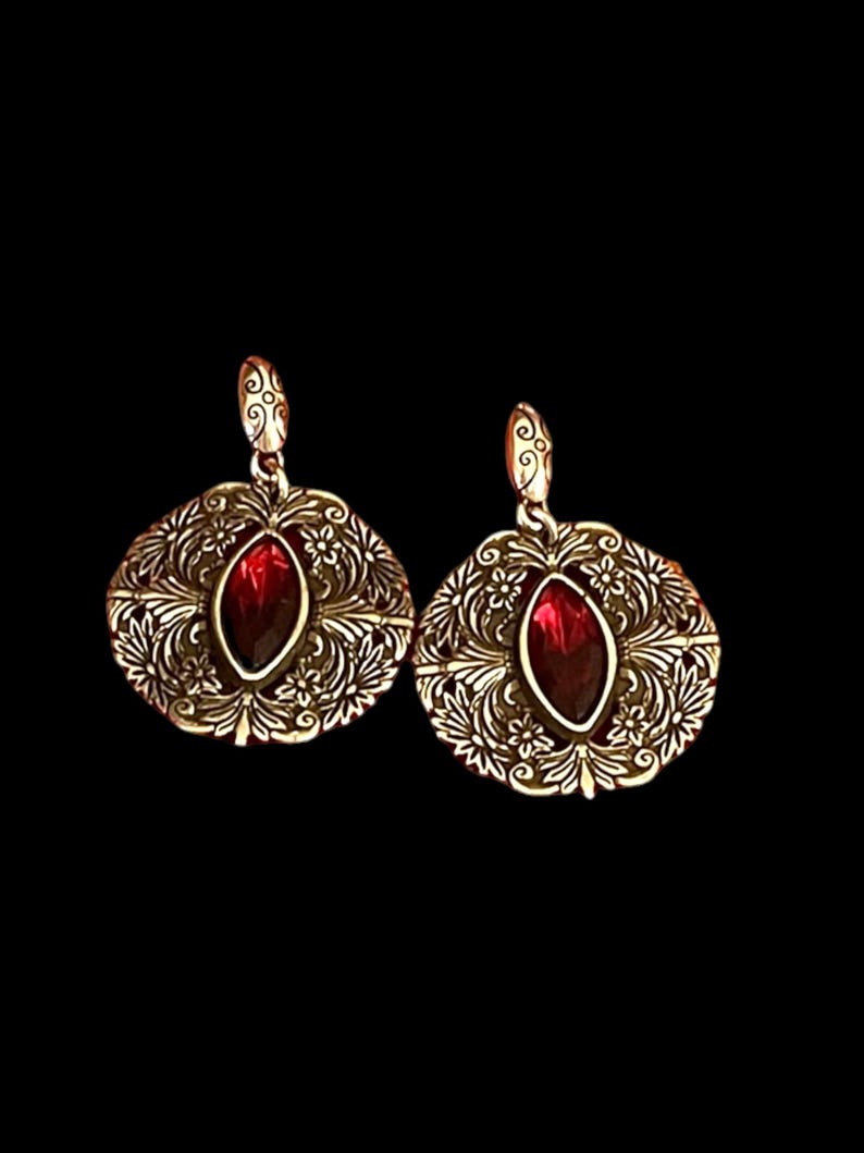 May include: A pair of gold-coloured earrings with a detailed filigree design. Each earring has a central, oval-shaped, deep red gemstone. The earrings feature a floral and scrollwork pattern around the gemstone.