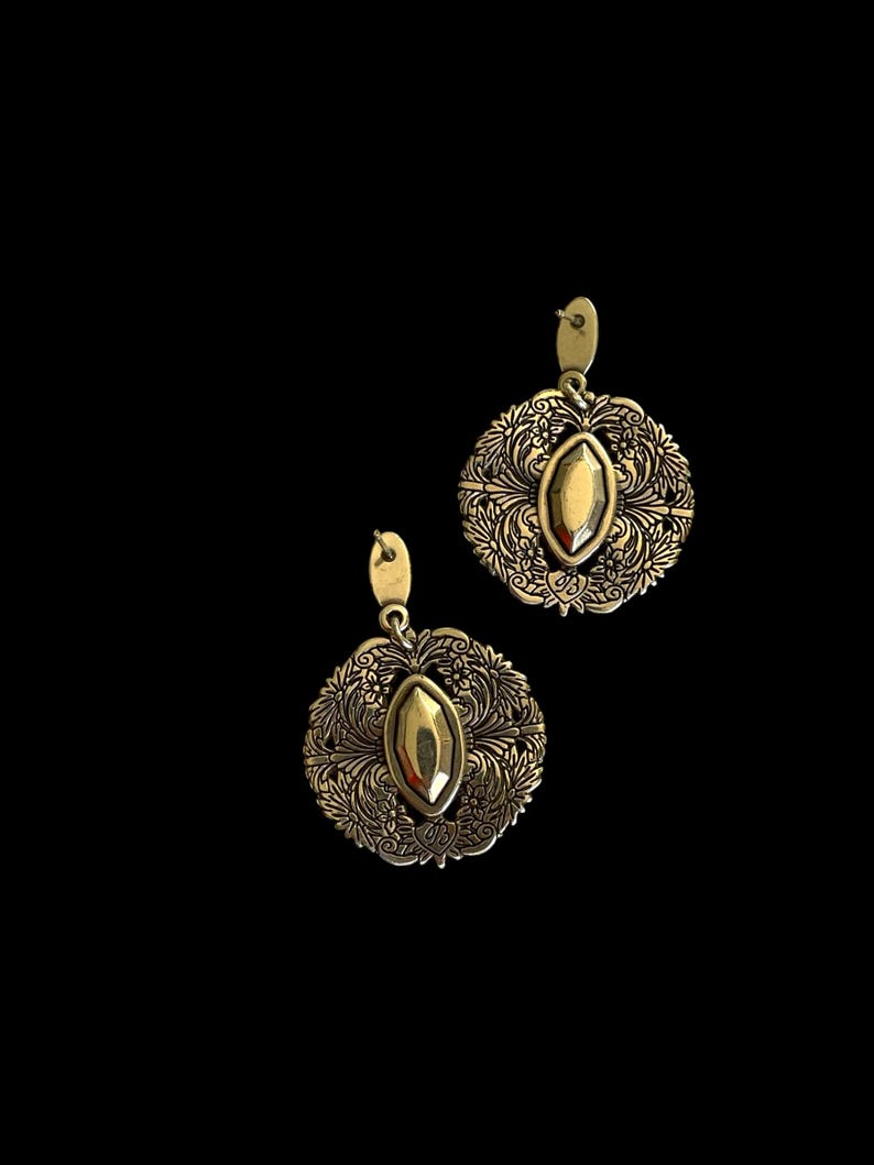 May include: A pair of gold-coloured earrings. Each earring has an oval-shaped stone at the centre, surrounded by a detailed floral design. The earrings have a hook closure and are set against a black background.