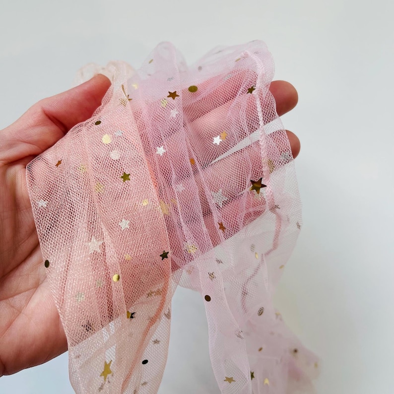 Puede incluir: Tela de tul rosa con estrellas doradas de purpurina. El tul es transparente y tiene una textura delicada.