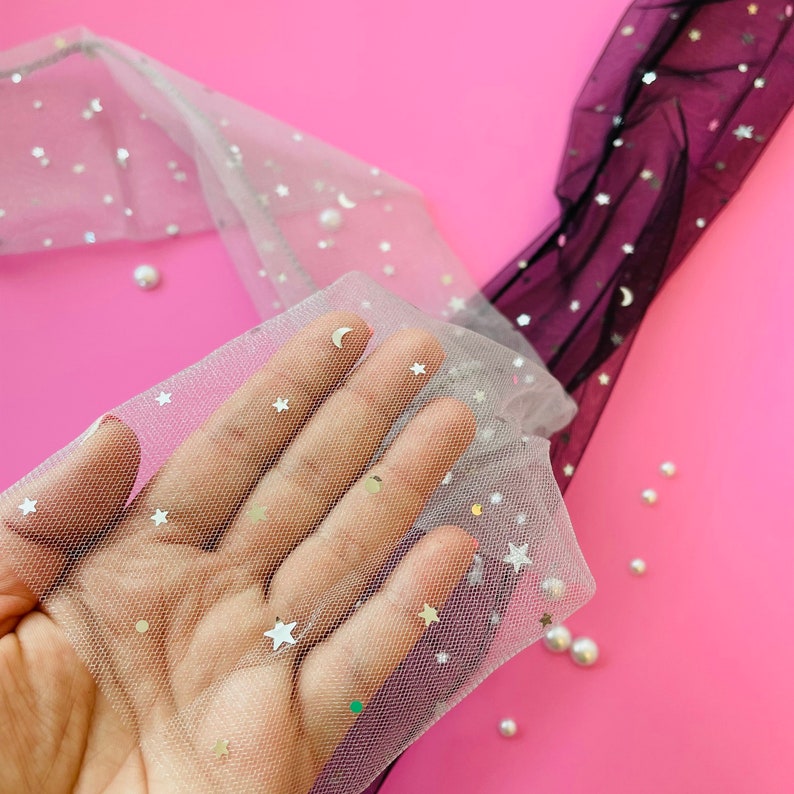 Puede incluir: Tela blanca transparente con estrellas y lunas plateadas brillantes. La tela es delicada y tiene una textura suave y fluida.