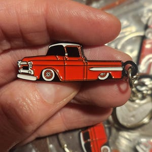 May include: A red and white enamel keychain featuring a classic pickup truck design. The truck has a white roof, white accents, and black tires. The keychain is attached to a silver-colored ring.