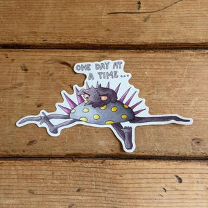 May include: A sticker featuring a cartoon creature with a gray body, yellow spots, and purple spikes. The creature is lying down with its arms and legs outstretched. The text "ONE DAY AT A TIME..." is above the creature.