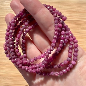 Könnte beinhalten: Mehrere Stränge facettierter, tiefrosa Edelsteinperlen werden gezeigt. Die Perlen sind zu mehreren Armbändern aufgereiht. Die Edelsteine haben ein funkelndes, poliertes Aussehen. Der Hintergrund ist eine helle Holzoberfläche.