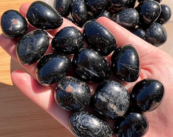 Può includere: Una collezione di pietre di tormalina nera levigate con una finitura liscia e lucida. Questi cristalli scuri e arrotondati presentano variazioni naturali nella consistenza e nella lucentezza, mostrando toni neri profondi con sottili riflessi.